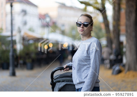Stylish young woman wearing sunglasses walking through Ljubljana city center pushing and rocking baby stroller. Warm autumn or spring weather for outdoor activity. 95927891