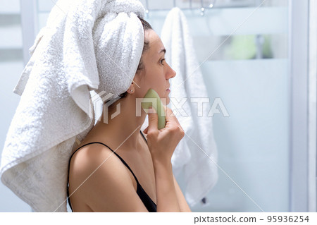 Woman makes a facial massage with a gouache scraper and looks in the mirror. She is wearing a nightie, towel on the head and has bare shoulders Woman makes a facial massage with a gouache scraper and looks in the mirror. She is wearing a nightie, towel on the head and has bare shoulders 95936254
