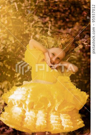 A little girl in a fluffy yellow dress. Little princess with blue eyes among the leaves and bushes 95937012