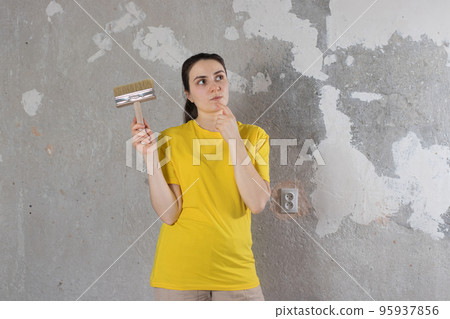 A woman repairman in a yellow bright t-shirt holds a brush and looks at an empty wall with place for text. Turnkey repairs 95937856