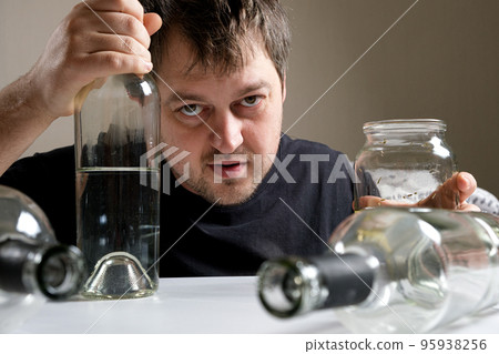 An alcoholic man sits at a table, holds a bottle of vodka with his hand and looks into the camera. Alcohol dependence, alcoholism and delirium An alcoholic man sits at a table, holds a bottle of vodka with his hand and looks into the camera. Alcohol dependence, alcoholism and delirium 95938256