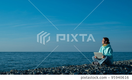 Happy caucasian woman working on a laptop while sitting on a pebble beach. Happy caucasian woman working on a laptop while sitting on a pebble beach. 95940616