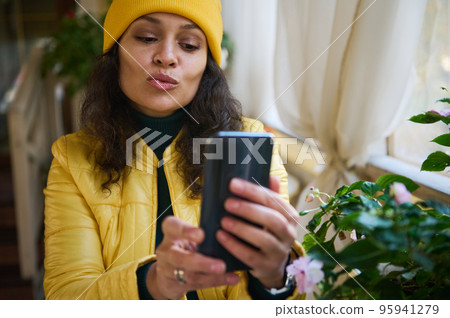 Beautiful multiethnic young woman, dressed in bright yellow jacket and hat, blowing an air kiss while talking by video conference on her mobile phone, sitting by table in a cafe during a coffee break Beautiful multiethnic young woman, dressed in bright yellow jacket and hat, blowing an air kiss while talking by video conference on her mobile phone, sitting by table in a cafe during a coffee break 95941279
