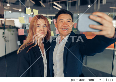 Two multi ethnic business colleagues taking selfie while standing in modern office Two multi ethnic business colleagues taking selfie while standing in modern office 95941444