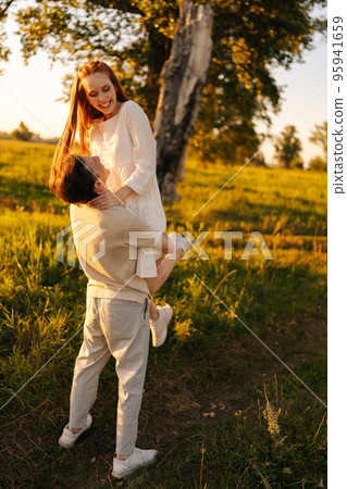 Vertical portrait of unrecognizable loving young man holding cheerful redhead woman in arms on green meadow in sunny summer evening, during golden sunset with soft sunlight. 95941659