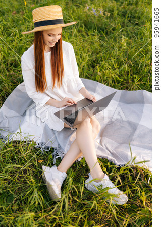 Vertical portrait of young female student in straw hat and white dress working on laptop computer sitting on beautiful field of green grass. Cute lady typing notebook looking to display outdoors. Vertical portrait of young female student in straw hat and white dress working on laptop computer sitting on beautiful field of green grass. Cute lady typing notebook looking to display outdoors. 95941665