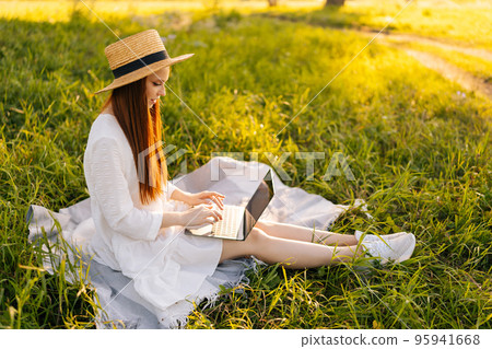 Side view of beautiful redhead woman in straw hat working with laptop sitting on green grass in summer park. Happy female student in white dress learning and surfing net, preparing for exams. 95941668