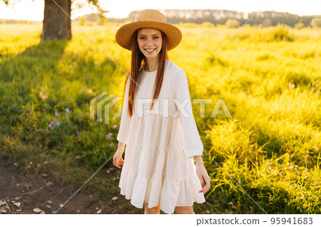 Portrait of cheerful redhead young woman in straw hat and white dress standing posing on beautiful field of green grass looking at camera, in sunset sunlight on summer evening. Portrait of cheerful redhead young woman in straw hat and white dress standing posing on beautiful field of green grass looking at camera, in sunset sunlight on summer evening. 95941683