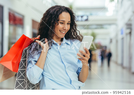 Happy and successful woman shopping for clothes in a supermarket store, Hispanic woman holding a smartphone reading online messages and browsing offers with discounts and sales Happy and successful woman shopping for clothes in a supermarket store, Hispanic woman holding a smartphone reading online messages and browsing offers with discounts and sales 95942516