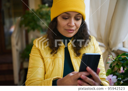 Close-up portrait of an African American cute woman dressed in bright yellow jacket and hat, relaxing in a coffee shop in warm autumn day, using phone, checking newsfeed and posting on social networks 95942523