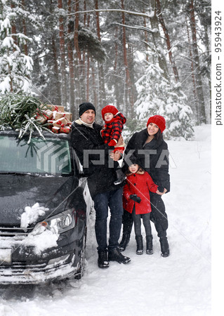 Mom and dad with their daughters in knitted red hats are standing by the car with a submerged Christmas tree on the roof. A trip to the forest for a Christmas tree 95943924