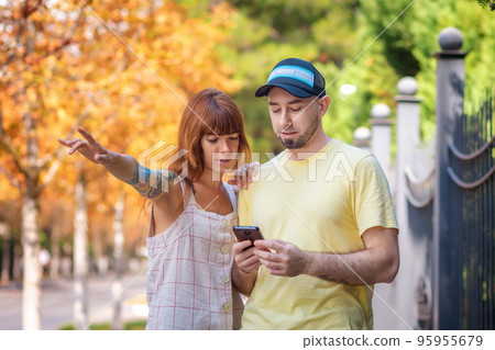 Young couple using a smartphone app with online map. Caucasian woman with tattoos shows a direction. The concept of relationship psychology and city travel Young couple using a smartphone app with online map. Caucasian woman with tattoos shows a direction. The concept of relationship psychology and city travel 95955679