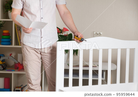 The furniture assembler assembles a children's white wooden bed, studies the assembly instructions 95959644