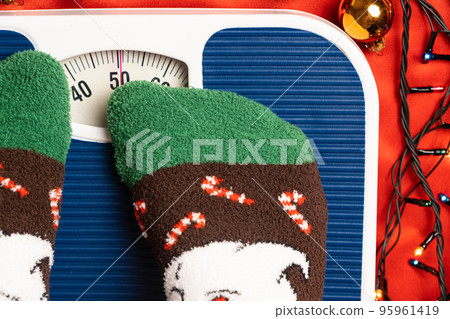 A woman in Christmas socks stands on the scales, weighs herself after gluttony during the holidays A woman in Christmas socks stands on the scales, weighs herself after gluttony during the holidays 95961419