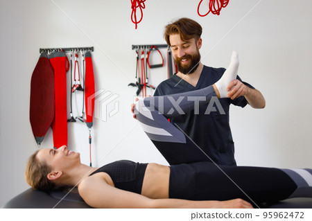 Side view of a cheerful physiotherapist doctor and his female patient who has problems with her leg. Rehabilitation specialist bending woman's knees Side view of a cheerful physiotherapist doctor and his female patient who has problems with her leg. Rehabilitation specialist bending woman's knees 95962472
