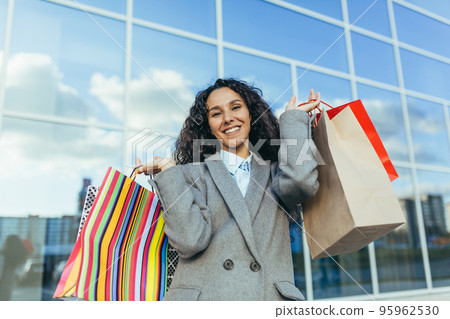 Beautiful woman with curly hair outside big store looking at camera and smiling, Hispanic woman shopper happy with discounts and sale, in warm coat portrait. Beautiful woman with curly hair outside big store looking at camera and smiling, Hispanic woman shopper happy with discounts and sale, in warm coat portrait. 95962530