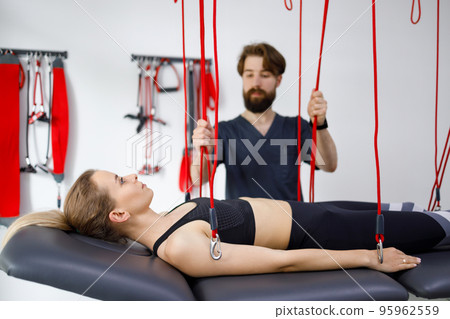 Physiotherapist assists female patient undergoing rehabilitative physiotherapy on a suspension rope system. Modern clinic using advanced rehabilitating therapy Physiotherapist assists female patient undergoing rehabilitative physiotherapy on a suspension rope system. Modern clinic using advanced rehabilitating therapy 95962559