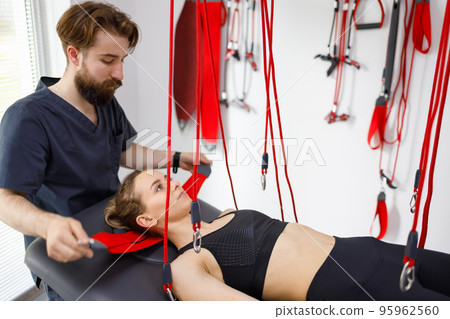 Physiotherapist assists female patient undergoing rehabilitative physiotherapy on a suspension rope system. Modern clinic using advanced rehabilitating therapy Physiotherapist assists female patient undergoing rehabilitative physiotherapy on a suspension rope system. Modern clinic using advanced rehabilitating therapy 95962560
