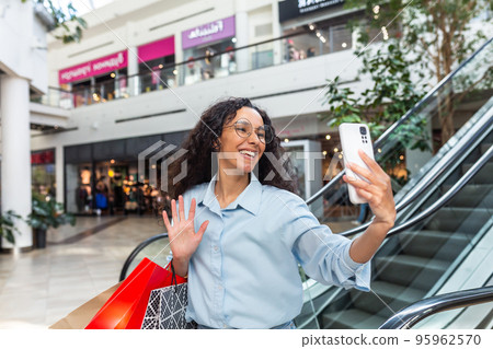 Happy female shopper inside store talking on video call with friends, Hispanic woman using smartphone for online remote communication, sharing information about sales and discounts. Happy female shopper inside store talking on video call with friends, Hispanic woman using smartphone for online remote communication, sharing information about sales and discounts. 95962570