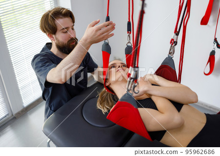 Physiotherapist assists female patient undergoing rehabilitative physiotherapy on a suspension rope system. Modern clinic using advanced rehabilitating therapy 95962686