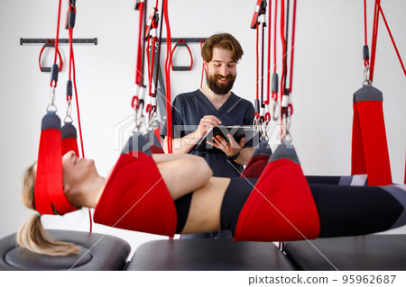 Physiotherapist doctor with clipboard watching female patient hanging on suspension system for neuromuscular activation in rehabilitation clinic Physiotherapist doctor with clipboard watching female patient hanging on suspension system for neuromuscular activation in rehabilitation clinic 95962687