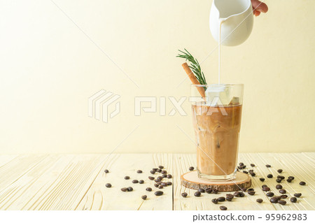 pouring milk in black coffee glass with ice cube, cinnamon and rosemary 95962983