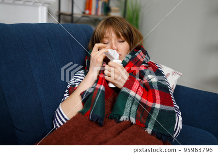 Sick woman with a runny nose lying on a sofa under a blanket with a handkerchief 95963796