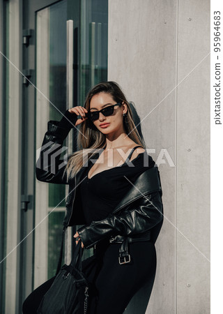 Fashionable young woman with long hair wearing solid long sleeve bodycon one piece jumpsuits posing in a black leather jacket and sunglasses Fashionable young woman with long hair wearing solid long sleeve bodycon one piece jumpsuits posing in a black leather jacket and sunglasses 95964683