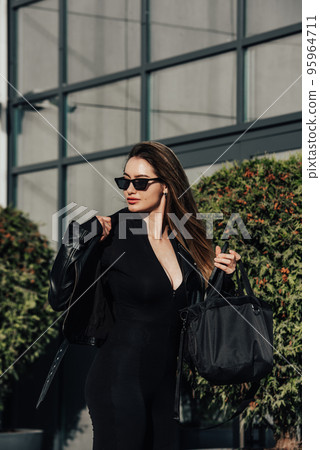 Fashionable young woman with long hair wearing solid long sleeve bodycon one piece jumpsuits posing in a black leather jacket and sunglasses Fashionable young woman with long hair wearing solid long sleeve bodycon one piece jumpsuits posing in a black leather jacket and sunglasses 95964711