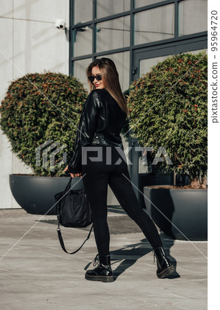 Fashionable young woman with long hair wearing solid long sleeve bodycon one piece jumpsuits posing in a black leather jacket and smooth platform boots Fashionable young woman with long hair wearing solid long sleeve bodycon one piece jumpsuits posing in a black leather jacket and smooth platform boots 95964720