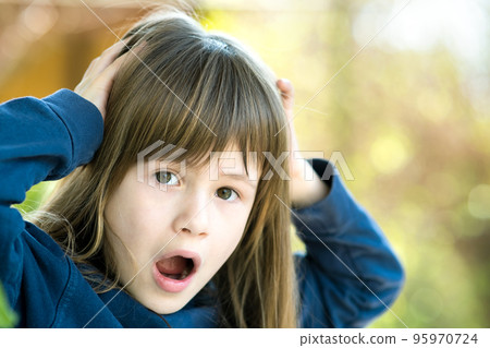 Portrait of surprised child girl holding hands to her head outdoors in summer. Shocked female kid on a warm day outside. Portrait of surprised child girl holding hands to her head outdoors in summer. Shocked female kid on a warm day outside. 95970724