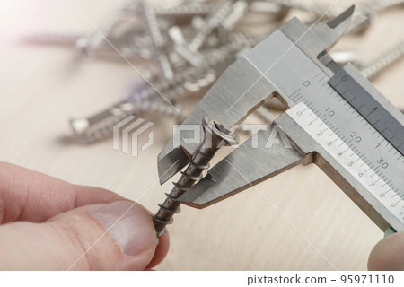 A man uses a caliper to measure the thickness of a screw with torx head. An accurate measuring device in the hands of a person against the background of screws scattered on the table A man uses a caliper to measure the thickness of a screw with torx head. An accurate measuring device in the hands of a person against the background of screws scattered on the table 95971110