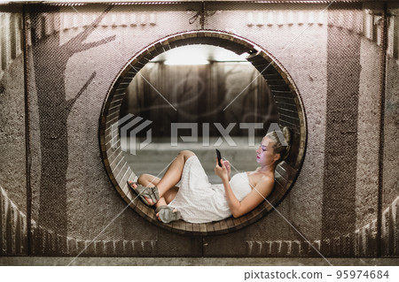 Lady wearing white summer dress playing carelessly with her android smart phone browsing trough social networks relaxing on contemporary circular bench in urban pedestrian underpass. 95974684
