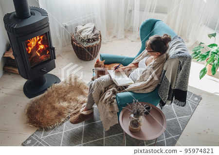 Young woman write in a notebook sitting in a armchair by the fi Young woman write in a notebook sitting in a armchair by the fi 95974821