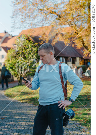Professional Photographer Taking Picture Of Beautiful Autumn Park. Man Professional Photographer standing With Camera And With Smartphone In Autumn Park. Retouched, Vibrant Colors Professional Photographer Taking Picture Of Beautiful Autumn Park. Man Professional Photographer standing With Camera And With Smartphone In Autumn Park. Retouched, Vibrant Colors 95988176