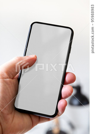 Hand holding a smartphone with blank grey screen. Office background. 95988983