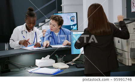 Multiethnic medical team analyzing checkup forms at registration desk in hospital reception lobby. Specialists and receptionist working on appointments at help counter. Handheld shot. 95991746