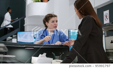 Portrait of medical assistant talking to receptionist at hospital registration counter, using appointment reports to help patients with checkup visits. Working at reception desk. Handheld shot. Portrait of medical assistant talking to receptionist at hospital registration counter, using appointment reports to help patients with checkup visits. Working at reception desk. Handheld shot. 95991797