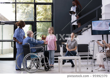 Granddaughter handing bouquet of flowers to disabled grandfather after finishing medical consultation in hospital waiting area. Elderly man discussing health care treatment with nurse Granddaughter handing bouquet of flowers to disabled grandfather after finishing medical consultation in hospital waiting area. Elderly man discussing health care treatment with nurse 95992370