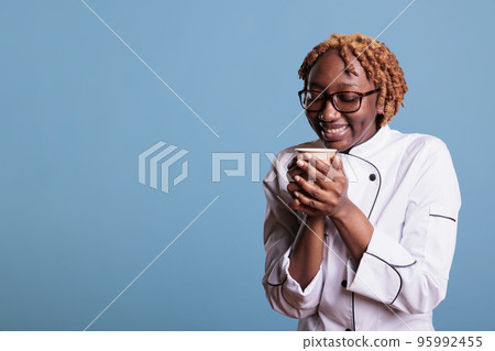 Relaxed female cook enjoying a cup of favorite coffee during lunch time. African american professional chef wearing kitchen uniform against blue background in studio shot. 95992455