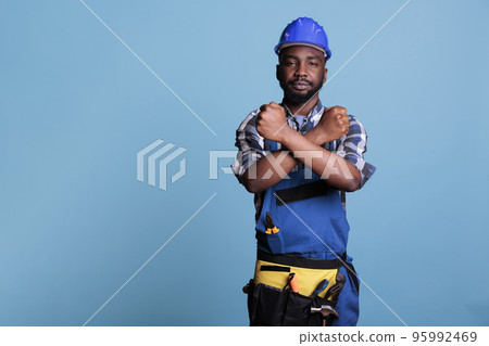 Builder making gesture of declining crossed arms while looking at camera. African american construction worker in work uniform giving declining hands symbol while standing on blue background. Builder making gesture of declining crossed arms while looking at camera. African american construction worker in work uniform giving declining hands symbol while standing on blue background. 95992469