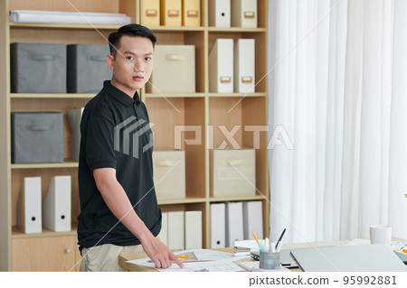 Serious young architecture bureau worker standing at desk with documents and blueprints Serious young architecture bureau worker standing at desk with documents and blueprints 95992881