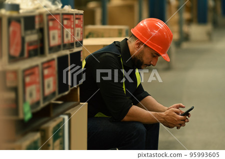 Side view of male worker sitting near cardboard boxes of goods and using mobile phone during taking a break Side view of male worker sitting near cardboard boxes of goods and using mobile phone during taking a break 95993605
