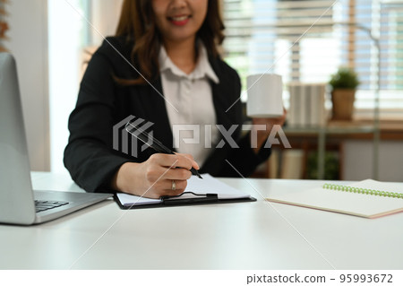 Smiling asian woman employee drinking coffee and working with some document at her office desk 95993672