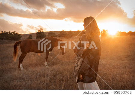 A girl in a white airy dress stands next to a horse and poses in the forest at sunset, like in a fairy tale. Well-groomed face with beautiful cosmetics. Enjoy nature and animals. Horse riding in the 95993762