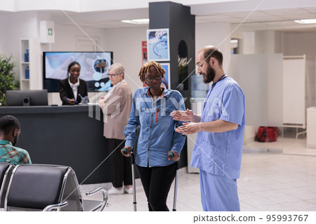 Hospital administrative staff guiding african american patient with degenerative disease to therapist office. Afro female receptionist at sanatorium supporting elderly woman. 95993767
