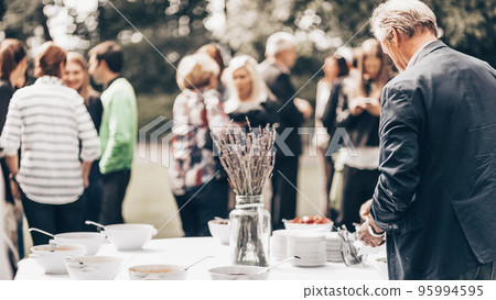 Banquet lunch break at conference meeting on hotel terrace. Assortment of food and beverage. Unrecognizable people discussing over food in background. 95994595