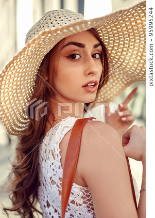Fashionable brunette woman in a white dress, straw hat, walking in the street, posing near shop windows 95995244