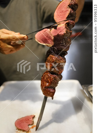 Picanha is a BBQ Steak meat grilled in charcoal. Knife cutting on skewer. Brazilian meat in a Churrascaria restaurant 95996313