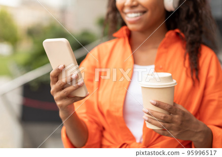 Cheerful millennial black lady in wireless headphones typing on phone, chatting, reading blog, enjoy walk in city Cheerful millennial black lady in wireless headphones typing on phone, chatting, reading blog, enjoy walk in city 95996517